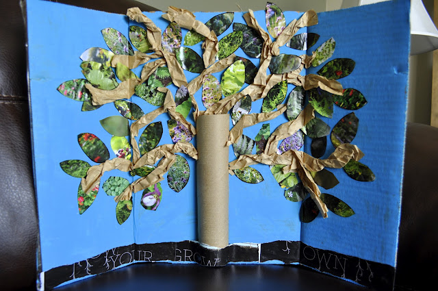 A creative 3D tree model made from a cardboard tube trunk and branches covered with crumpled brown paper. The leaves are crafted from cut-out magazine pieces, set against a blue cardboard backdrop.