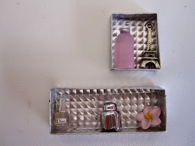 Two silver-coloured embossed metal dolls house wall boxes, one rectangular and one square, with a pink bottle and model Eiffel Tower in the top one and a perfume bottle, silver bottle and frangipani in the lower one.