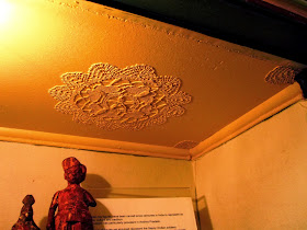 Ceiling of a room in the dolls house display cabinet, showing doileys stuck to it to replicate plaster moldings