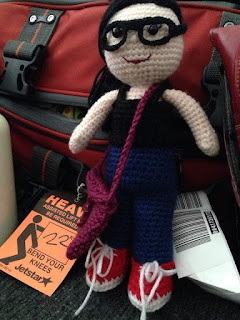 Kwokkie Doll is dressed in travelling clothes: blue jeans, red sneakers, black tank top and glasses. She has a purple satchel slung over her shoulder as she leans on the flight bag of the Little Colourful Teacher