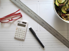 Reading glasses, metal ruler, calculator, pen and bowl of pasta on top of a piece of paper with a plan for a wall with windows.