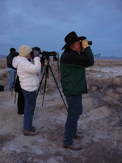 MAn and woman stand with binoculars looking off camera.
