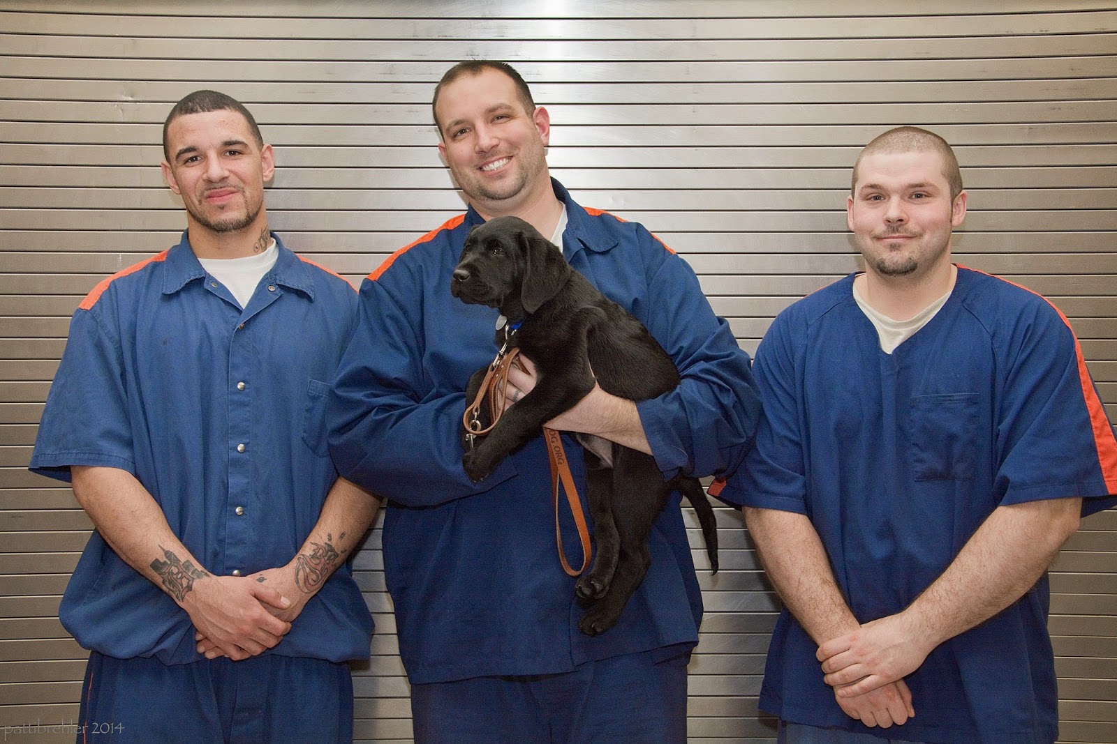 Three men are standing in front of a steel wall. They are wearing prison blue uniforms. The middle man is cradling a young black lab in his left arm with a leather leash hanging down. The men on either side have their hands folded in front of themselves. The men are all smiling at the camera.