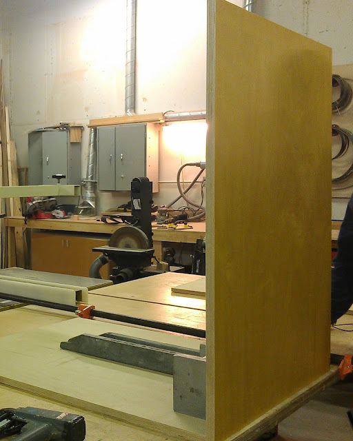 The casework starts coming together On a table sits to pieces of wood at a right angle. Each is yellow and measures 34 inches by 34 inches by 34 inches. There is a long metal weight along the piece lying parallel with the table to help support the one that is perpendicular to the table. There is a belt sander in the background which has a wheel about half way up a post that has the belt used for sanding on it. There are also grey cabinets along the walls.
