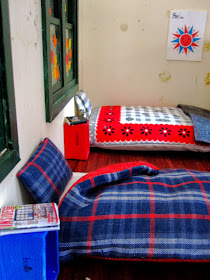 Miniature holiday house scene showing two mattresses on the floor, made up with sheets, pillows and doonas. Next to each is a wooden box on end with reading material on them.