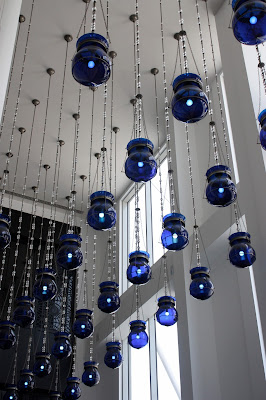 rows of blue lightbulbs hang from a high white ceiling in front of open windows