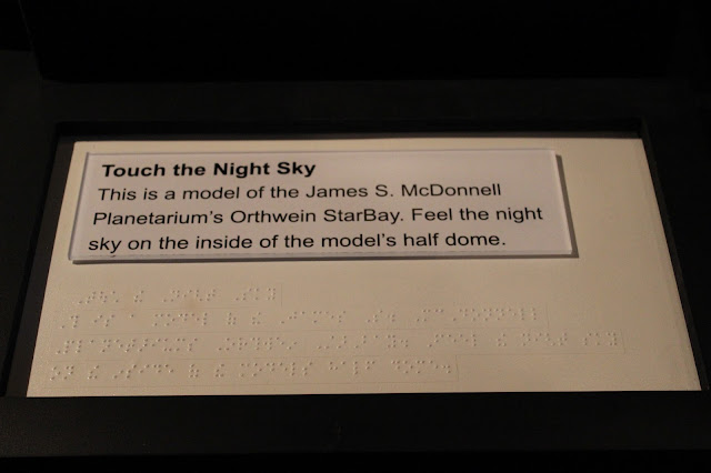 Exhibit text explaining the purpose of the model In large print and Braille reads the following exhibit signage: "Touch the Night Sky. This is a model of the James S. McDonnell Planetarium's Orthwein StarBay. Feel the night sky on the inside of the model's half dome."
