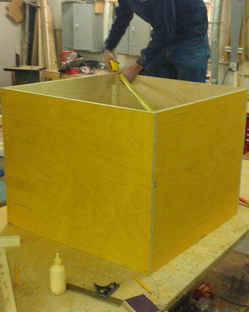 A box is formed On a table sits the casework. It is a yellow box with no top that measures 34" by 34" by 34". A young man in a long sleeved dark blue shirt stands behind it on the table measuring the diagonal distance between two points on the top of the box with a yellow tape measure. A bottle of yellow colored wood glue can be seen in front of the box and the grey cabinets can be seen on the wall in the background.