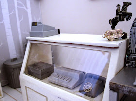 Counter of a modern dolls' house miniature homeware shop in grey and white. On the counter is a cash register and two silver statues. On display under the counter is a vintage grey typewriter and a vintage grey telephone.