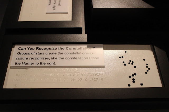 Exhibit text explaining constellations In large print and Braille reads the following exhibit signage: "Can You Recognize the Constellation? Groups of stars create the constellations our culture recognizes, like the constellation Orion the Hunter to the right." To the right of the text is a raised version of the constellation Orion like what would be found inside the dome made of black rivets on the white panel of text.
