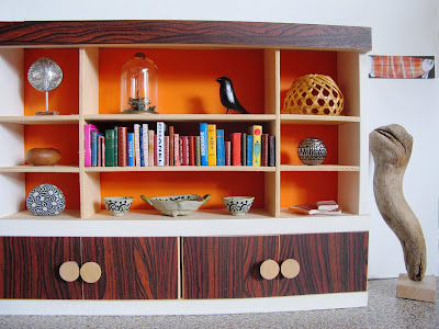 Modern dolls' house miniature scene of a 1970s display cabinet holding a selection of art objects and books. Next to it is a wooden sculpture, and an aboriginal artwork on the wall.