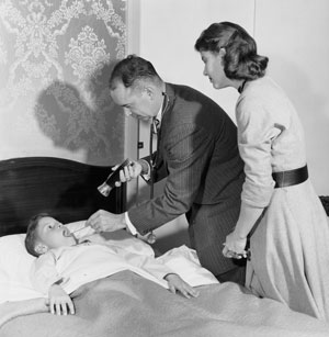 picture of a doctor and a lady looking down at a child in a bed. The doctor has a flashlight that he is using to shine line into the child's mouth so that he can see better.