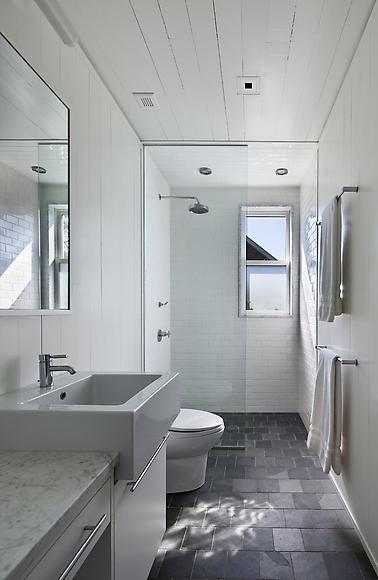 Classic meets modern in a bathroom with white paneled ceiling, flagstone tile floor, marble sink and counter top, drawers with long pulls, and shower with an overhead shower head, white tile backsplash and floor length glass door
