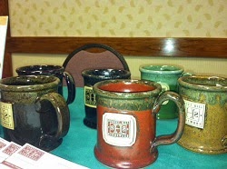 Custom made ceramic coffee mugs Table with green tablecloth displaying handmade ceramic coffee mugs painted brown, mustard, dark blue and green.