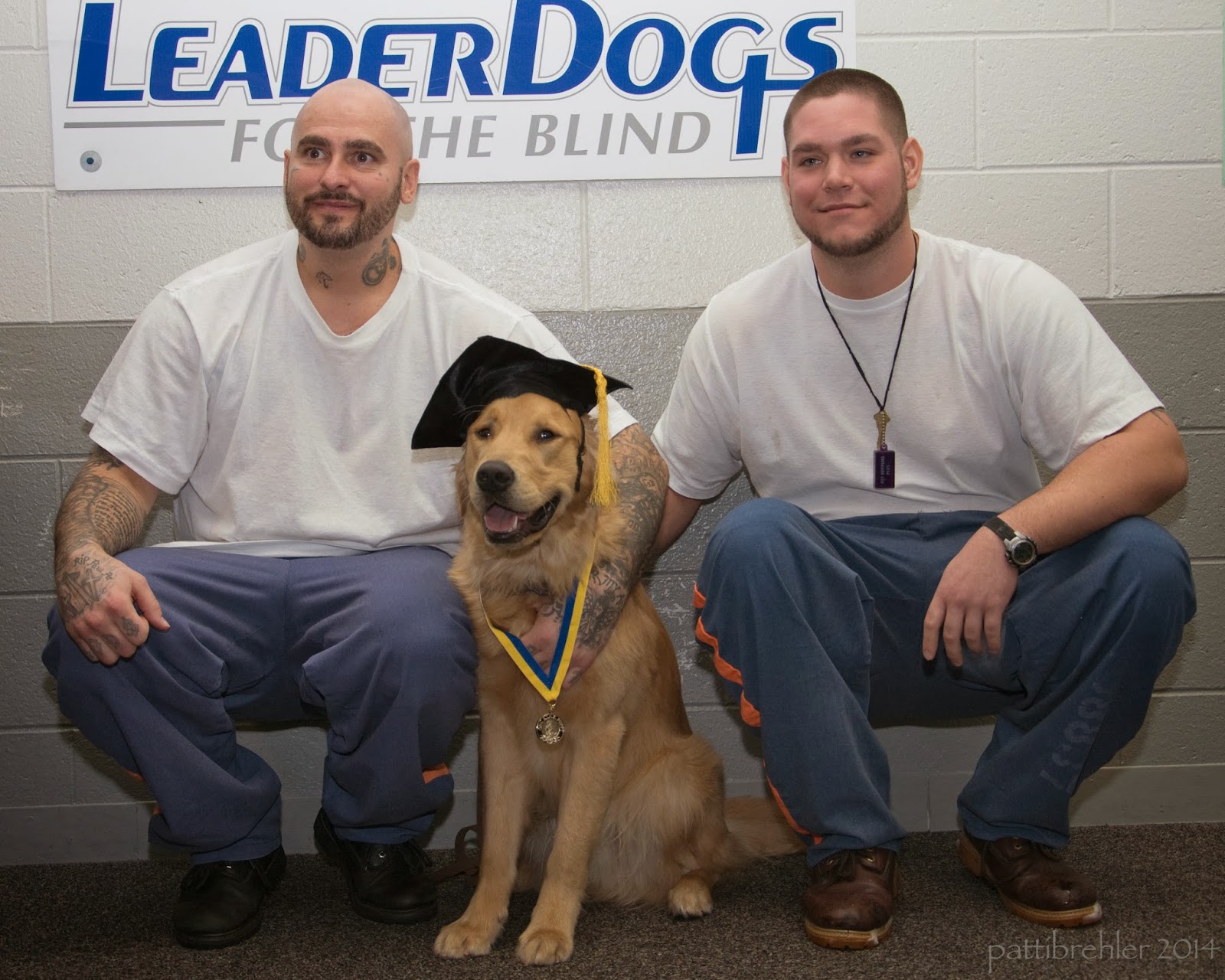 Two men are squatting down with a golden retriever sitting betweent hem. The men are wearing white t-shirts and blue pants. The golden retriever is wearing a black graduation cap and a yellow and blue ribbon with a medal around its neck. Behind them on the wall is a white poster with Leader Dogs for the Blind on it.