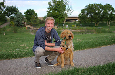 Ian smiles posing with a Golden Retriever guide dog puppy with a green field and trees behind them. Ian smiles posing with a Golden Retriever guide dog puppy with a green field and trees behind them.