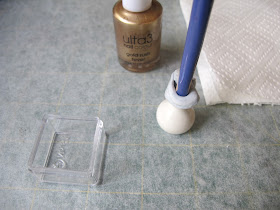 White dolls' house miniature vase, with the end of a paint brush blue tacked into it, on a piece of baking paper with a bottle of nail varnish, a small plastic container and a piece of paper towel.