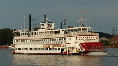 present day photo of the belle of louisville on the ohio river with steam coming out of the stacks with blue skys and green river and white and red painted boat