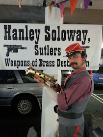 A man dressed in colonial attire, holding a steam-punk-style model gun, in front of a sign saying 'Hanley Soloway, Sutlers: Weapons of brass destruction'