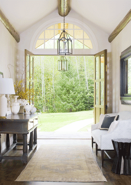 Foyer with double doors, crystal pendant light, a wood floor, exposed beams, a faded rug, a white sofa and a stained wood sidetable