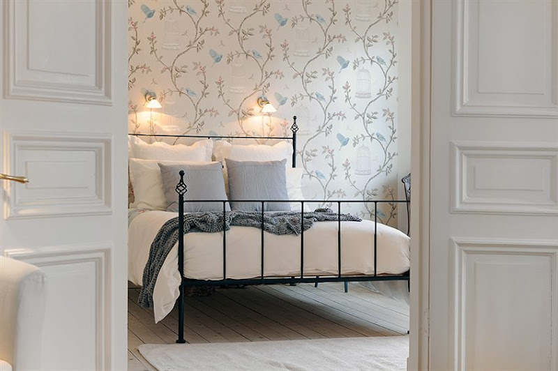 Bedroom in a Swedish apartment with iron bed frame, wall mounted lights, wall covered in a delicate wallpaper with a floral pattern with birdcages