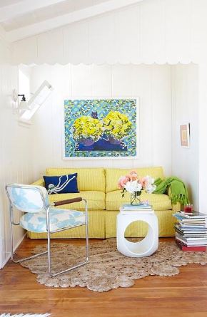 Small sitting room with a yellow sofa, white paneled walls, a wood floor, a stack of books being used as a side table, a white modern coffee table, a chair with metal arms and legs and a blue and white cushions and a crocheted rug