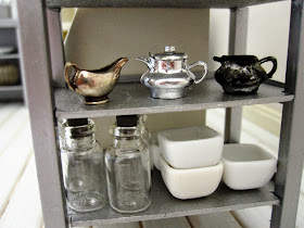 Close up of  a modern dolls' house miniature display of homeware. On the top shelf are three silver vintage jugs, and below are several glass bottles and white bowls.