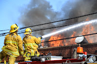 Two firefighters atop fire engine squirting water on top of a burning building. Copyright Photo by Ryan Babroff. Click to view more...