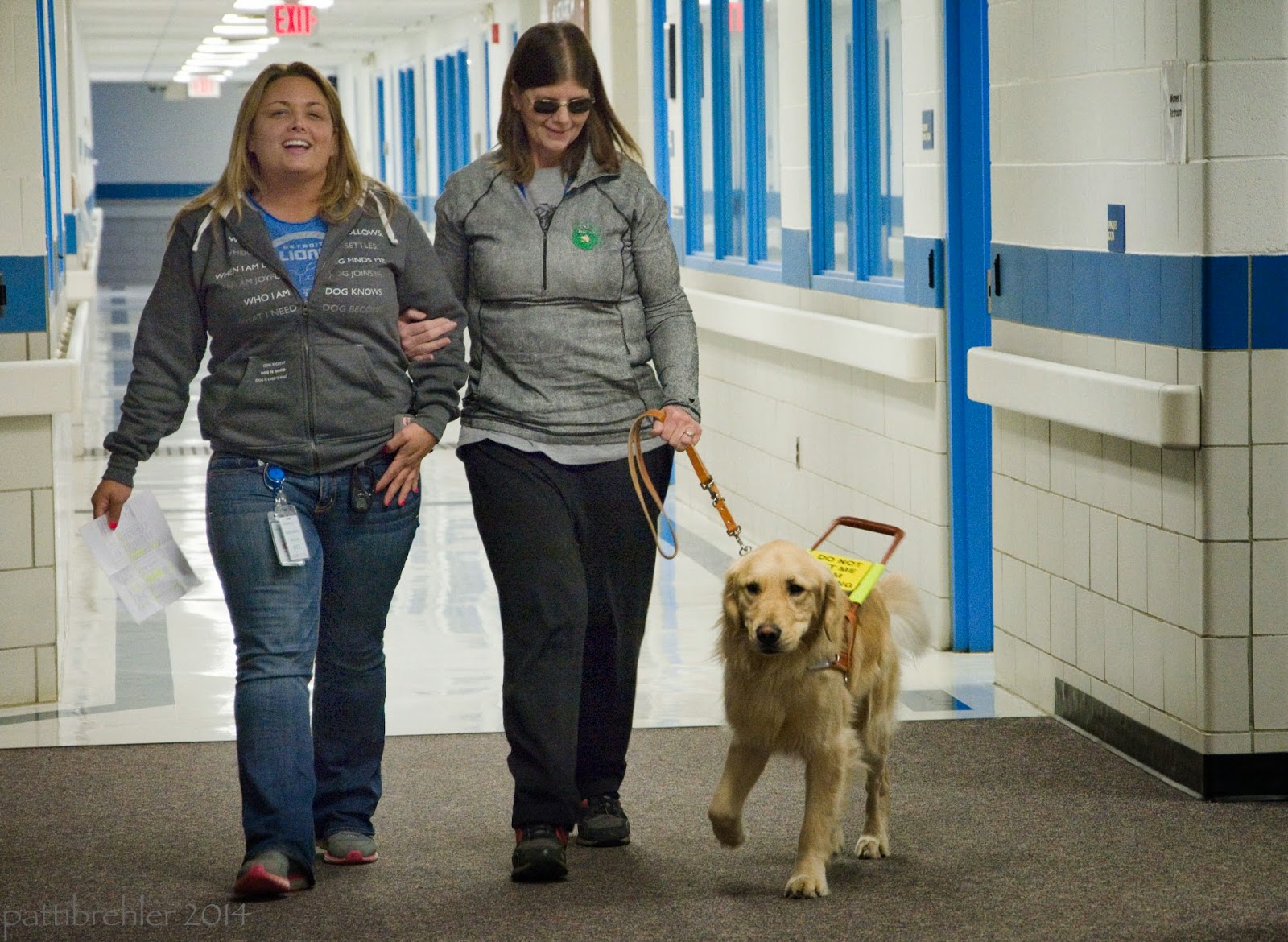 Two women are walking arm in arm toward the camera. The woman on the left is a Leader Dog trainer, she is wearing blue jeans and a grey zippered hoody and a blue t-shirt under it. Her ID tag is pinned to the hem of the sweatshirt. She has longish blond hair and she is smiling. The woman on the right has her right hand in the crook of the other woman's elbow. She is wearing black slacks and a grey longsleeved top. She is wearing sunglasses and has longish brown hair. She is holding a leash in her left hand that is attached to a golden retriever on her left side. The golden has a Leader Dog harness on with a yellow sign that says please don't pet me I'm working. The women and dog are just on a carpeted floor, behind them is a long hallway with tile floor.
