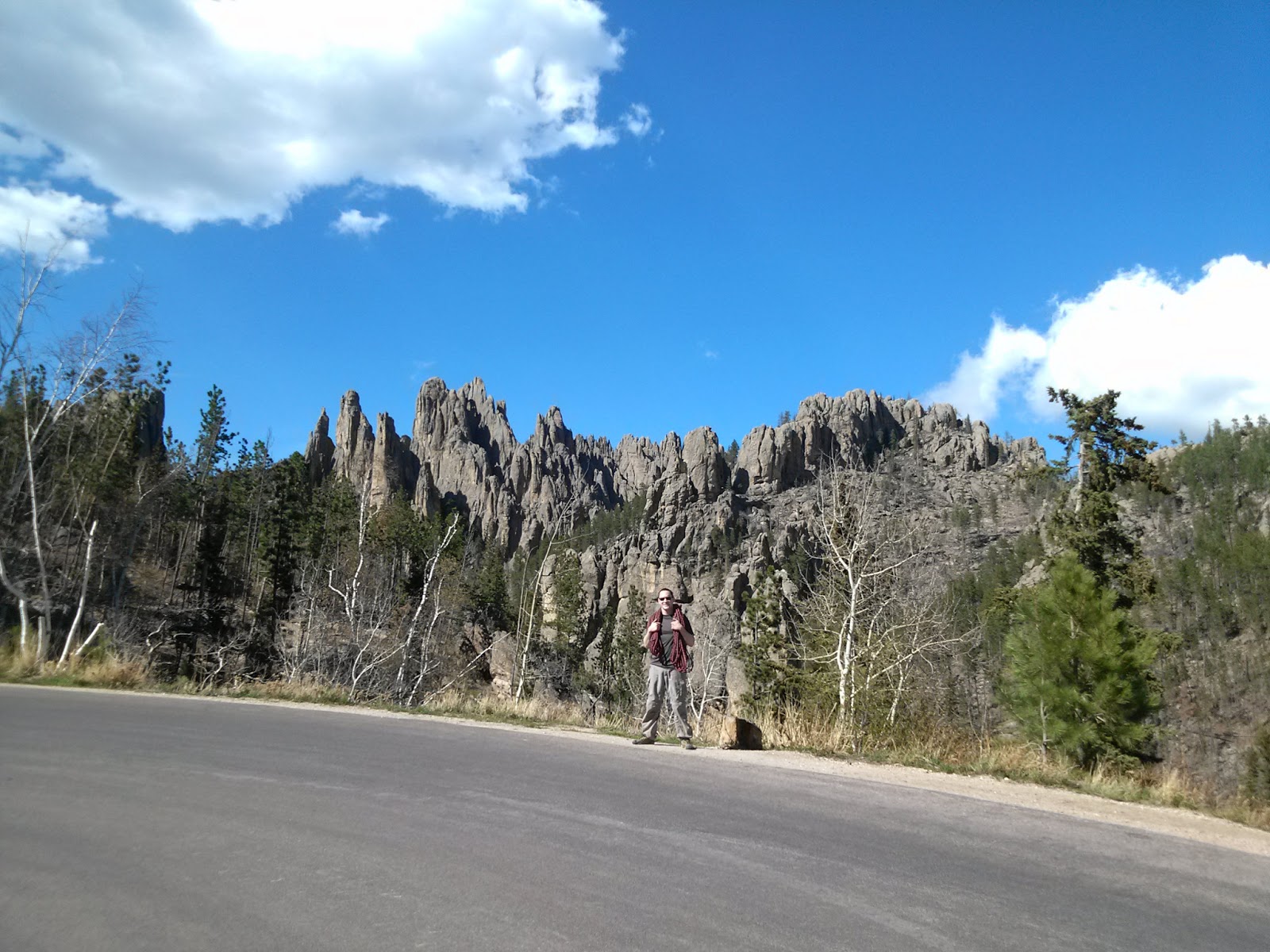 Man with a rope slung over his shoulders stands in front of the jagged Cathedral Spires against a bright blue sky after a day of guided climbing in Custer State Park.