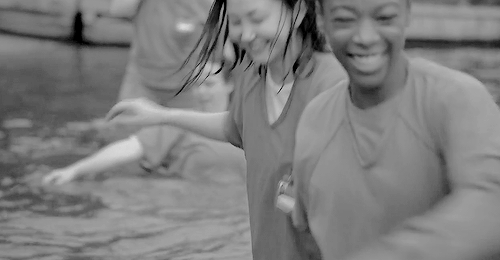 No gif: Duas meninas com uniforme de prisão molhadas pulando na água de um lago sorridentes com várias outras se divertindo no fundo.