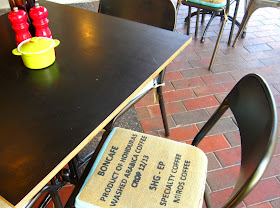 Black cafe table and chairs at Ona cafe, with red pepper grinder and green sugar bowl. The chair seat covers are made from coffee sacks.