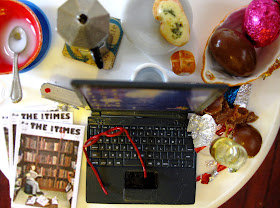 Modern dolls' house miniature round table with a laptop computer, digital camera, reading glasses, back issues of The tiny Times, and plates and bowls and cups containing half-eaten pieces of food, including Easter eggs.