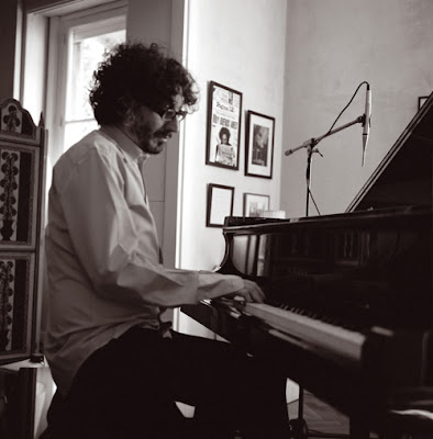 Foto em preto e branco mostra Fito Paez tocando um piano em uma pequena sala, com fotos emolduradas na parede, ao fundo. Foto em preto e branco mostra Fito Paez tocando um piano em uma pequena sala, com fotos emolduradas na parede, ao fundo.