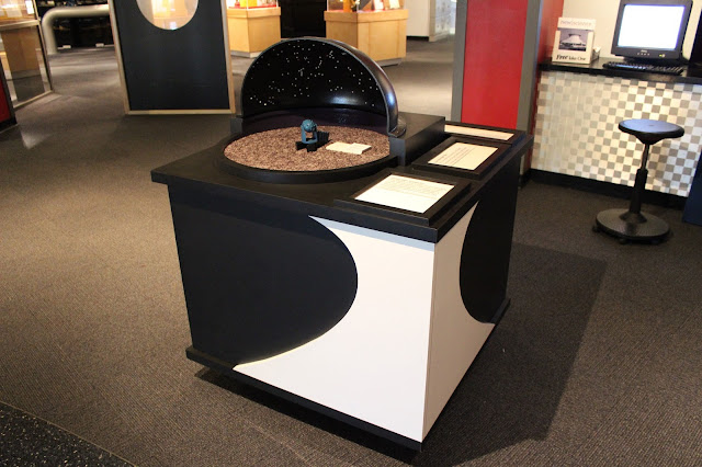 Waiting for its first visitors! The exhibit is now out in the Planetarium lobby. Other casework that is light brown can be seen behind it in the background as well as a black wall and a red pillar. The black box casework sits at about 34" by 34" by 34". On the front wrapping around the corner to the right side of the casework is the curved single sheet hyperboloid shape of the Planetarium in white. On the top of the casework is the black dome with all the stars of white rivets sits upon the purple wall and grey carpet. In the middle is the Zeiss model. The teal oval StarBall on stilts sits on the two smaller black wedges and the teal eight planet projector vertical boxes sit on the long black wedge. Next to it on the carpet sits a small white exhibit text panel of about three inches by one-and-a-half inches. It has Braille and large print marking the StarBall as exhibit one and the Planet Projectors as exhibit two to correspond to the other exhibit text. To the right of the model is the exhibit text. There are three visible rectangles of large print and Braille text framed by black.