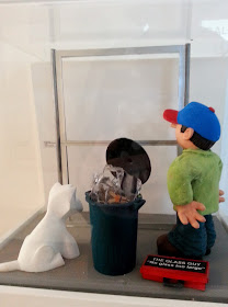 Miniature clay figure and dog, looking at a window space in a gallery exhibition.