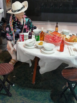 Miniature scene of a person at a table holding various plates of seafood and bottles.