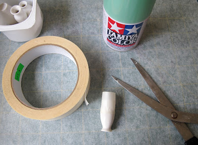 Container of dolls' house miniature white vases, a roll of masking tape, a pair of scissors and a can of spray paint arranged on a piece of baking paper.