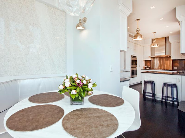 Breakfast nook and kitchen in an apartment with banquette seating around a white round table with a piece of modern art. The white kitchen has black counter tops, clear Kartell Lucite purple bar stools and two pendant lights