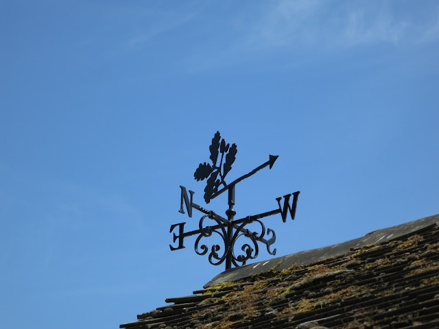 Metal weathervane with oak leaf symbol on top of tiled roof with moss.