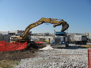 Workers use water to control dust as an excavating machine lifts soil into a dump truck. When hauled away for disposal, the soil will be covered.