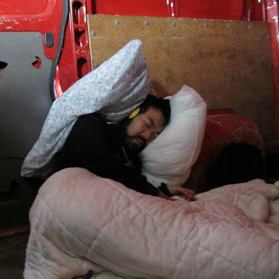 a student asleep in the back of a van, their head covered in pillows