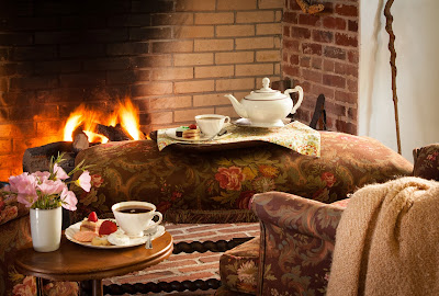 a tea pot and tea cup and saucer set on a bench in front of the fireplace and a plate with tea sandwiches and cookies on a table next to a chair nearby