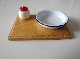 Dolls' house miniature chopping board with a enamelware bowl and red and cream jar on it.