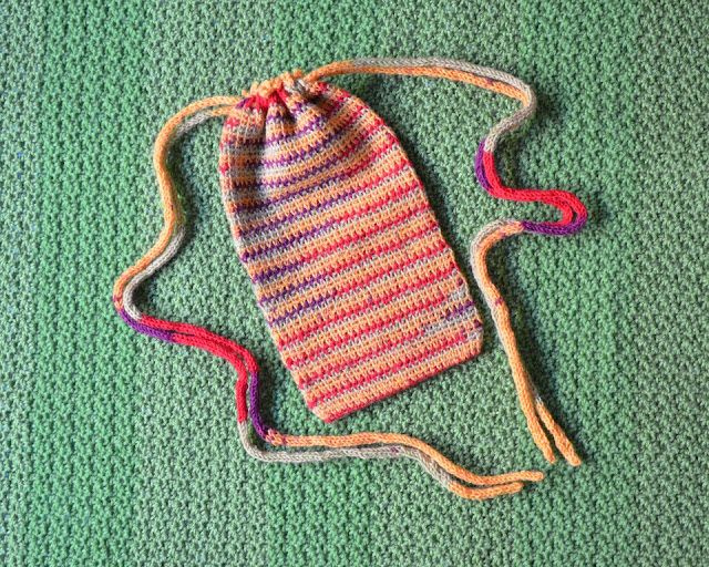 The striped bag is laying on a crocheted green background.  The drawstrings have been pulled which closes the bag by cinching it in at the top.