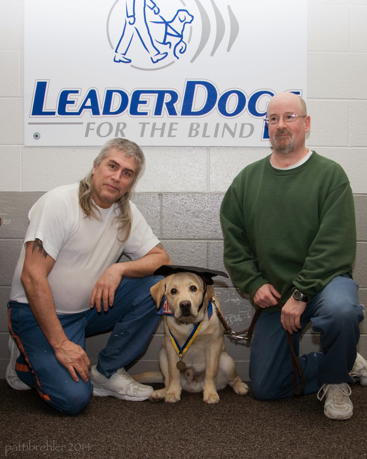 Two men are kneeling on one knee with a golden retriever between them. The man on the left is wearing a white t-shirt and blue pants. The man on the right is wearing a green sweatshirt and blue pants. Behind them on the wall is a white poster with Leader Dogs for the Blind written on it. The dog is wearing a black graduation cap and a yellow and lbue ribbon with medal around its neck.
