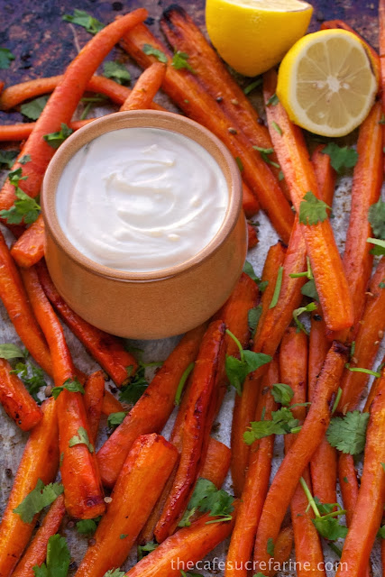 Honey Roasted Carrots - thecafesucrefarine.com A closeup photo of a pan of Honey Roasted Carrots with a dish of lemon tahini dressing in the center and lemon halves as a garnish.