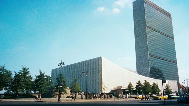 Image Description: Swooping white building in the foreground with curved roof, with slate-blue rectangular skyscraper in the background. 60s-era slightly grainy color image, vintage taxis are seen small in the foreground.