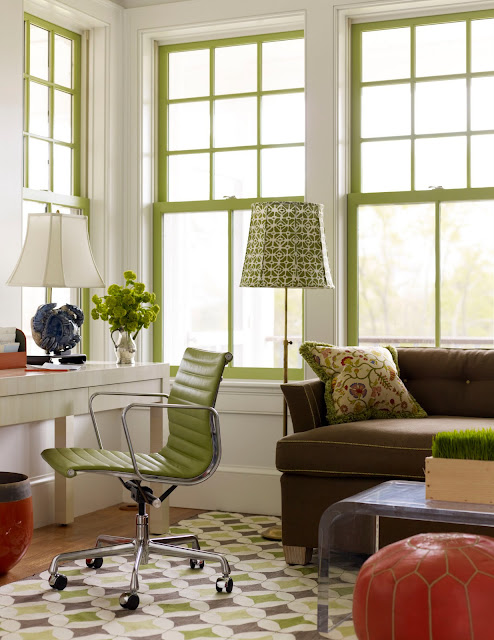 Home office with a brown sofa with chartreuse piping, a white desk with a metal rolling chair with a chartreuse seat and back, white walls, chartreuse window trimmings and an orange Moroccan pouf with chartreuse stitching and a Lucite coffee table 