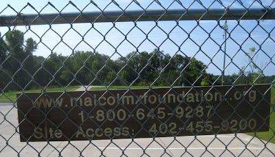 Chain link fence with parking lot visible through it, and sign giving the Malcolm X Foundation web address